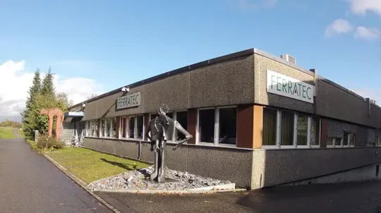 Industriegebäude mit Ferratec-Logo, grauer Fassade, großen Fenstern und Statuen im Vorgarten auf einem bewölkten Tag.