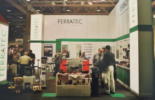 Messestand von Ferratec mit Besuchern, Technikprodukten und Informationsständen; Austausch zwischen zwei Männern im Vordergrund.