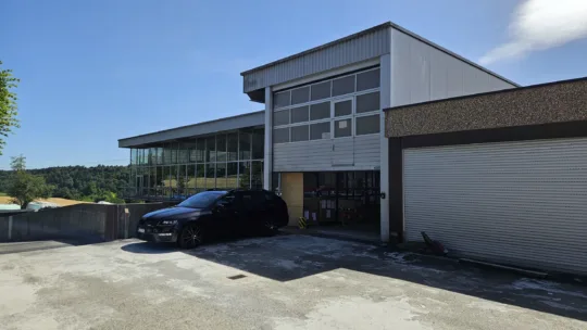 Industriegebäude mit verglaster Fassade, schwarzes Auto im Vordergrund, Lageröffnung erkennbar, umgeben von grüner Landschaft.