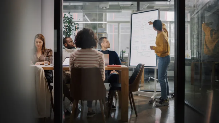 Person präsentiert Diagramm auf Whiteboard in modernem Büro; Teammitglieder am Tisch folgen aufmerksam der Präsentation.