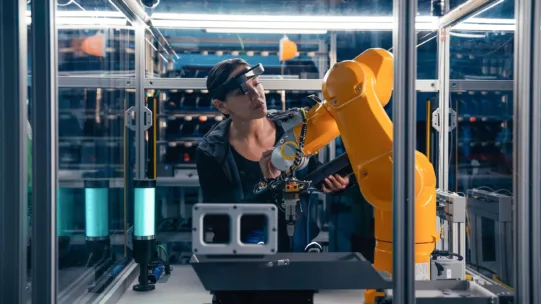 Ingenieurin überprüft in Hightech-Fabrik einen gelben Industrieroboterarm mit Spezialbrille zur präzisen Wartung.