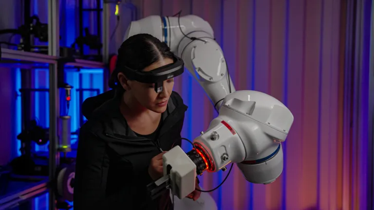 Frau mit AR-Brille arbeitet konzentriert an einem großen weißen Roboterarm in einem beleuchteten Labor.