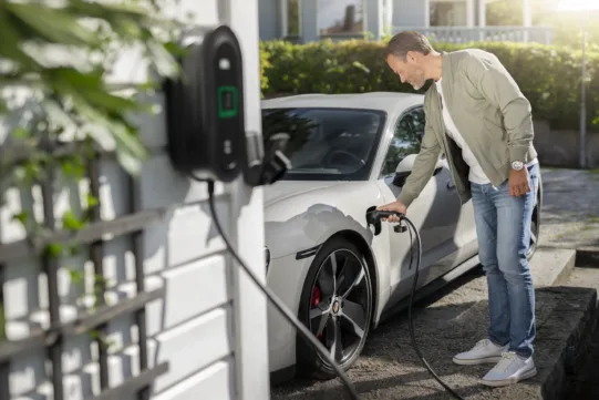 Mann lädt weißes Elektroauto an Wandladestation im Freien. Umweltfreundliche Mobilität, Ladekabel, moderne Technologie.
