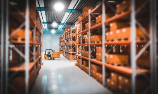 Gabelstapler transportiert orangefarbene Kiste in einem Lager mit hohen Regalen, beleuchtet von Industrielampen.