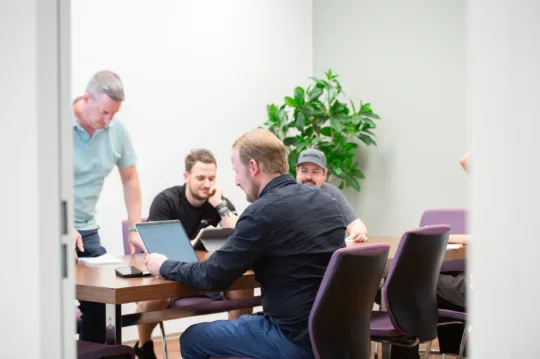 Menschen arbeiten am Konferenztisch mit Laptops und Tablets, umgeben von grüner Büropflanze, in modernem Büro.