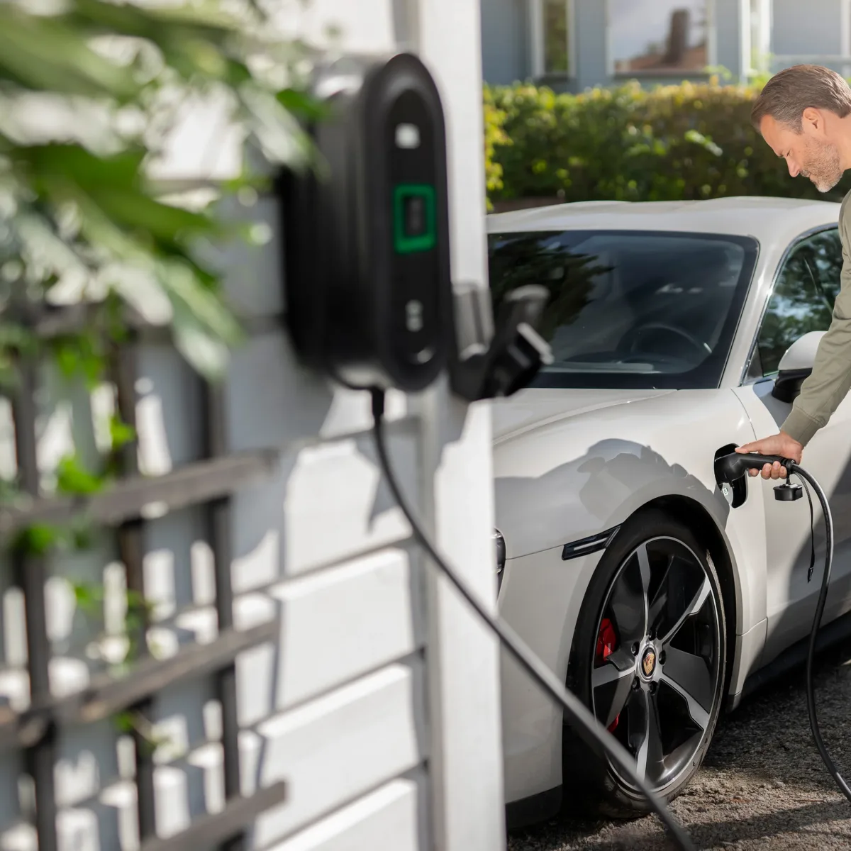Mann lädt weißes Elektroauto an einer Wallbox im Freien auf, Ladegerät-Kabel verbunden, umweltfreundliche Mobilität.