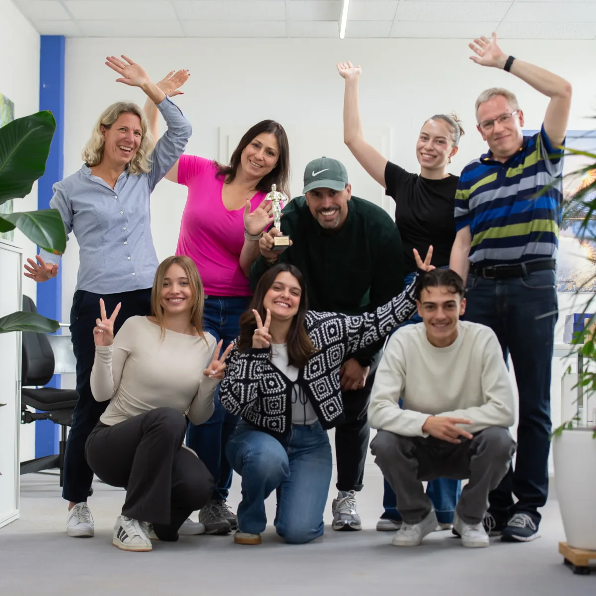 Lächelnde Gruppe von acht Personen posiert freudig in einem Büro mit Pflanzen, einer Person hält eine kleine Trophäe hoch.