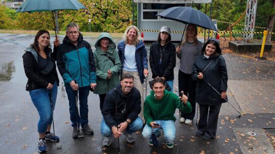 Gruppe von Menschen mit Regenschirmen und Gehhilfen posiert lächelnd auf nasser Straße im Herbst, Bäume im Hintergrund.