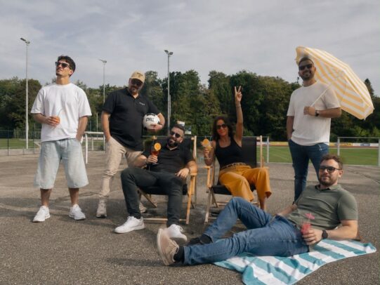 Gruppe von sechs Personen entspannt auf einem Sportplatz, sitzt auf Stühlen und Decke, mit Getränken und Sonnenschirm.
